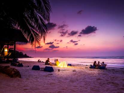 Young people sitting on a tropical beach at sunset, and the sky is filled with dark pinks and purples.