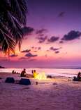 Young people sitting on a tropical beach at sunset, and the sky is filled with dark pinks and purples.